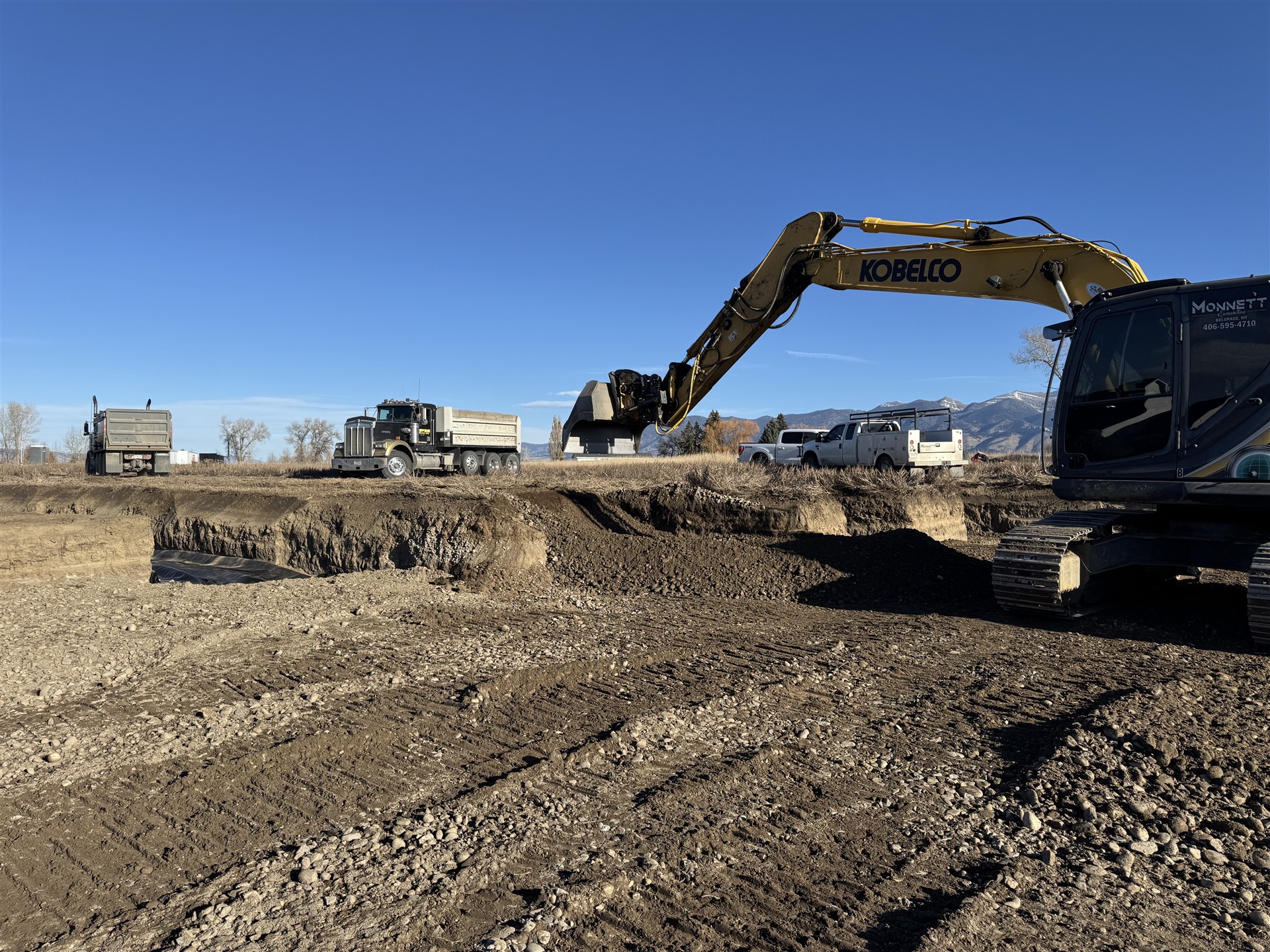 Monnett excavator and gravel trucks at an excavation site with Montana mountains in the distance